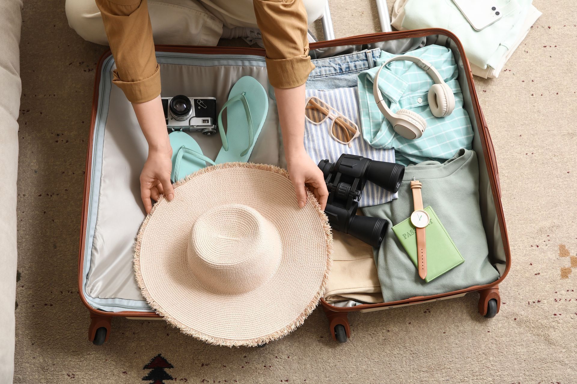 Person packing a suitcase with sandals, clothes, camera, and a straw hat on a beige floor
