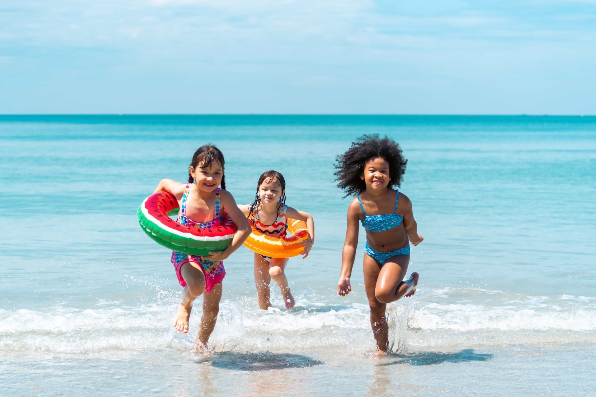 Three children in colorful swim rings running through shallow ocean waves on a sunny beach