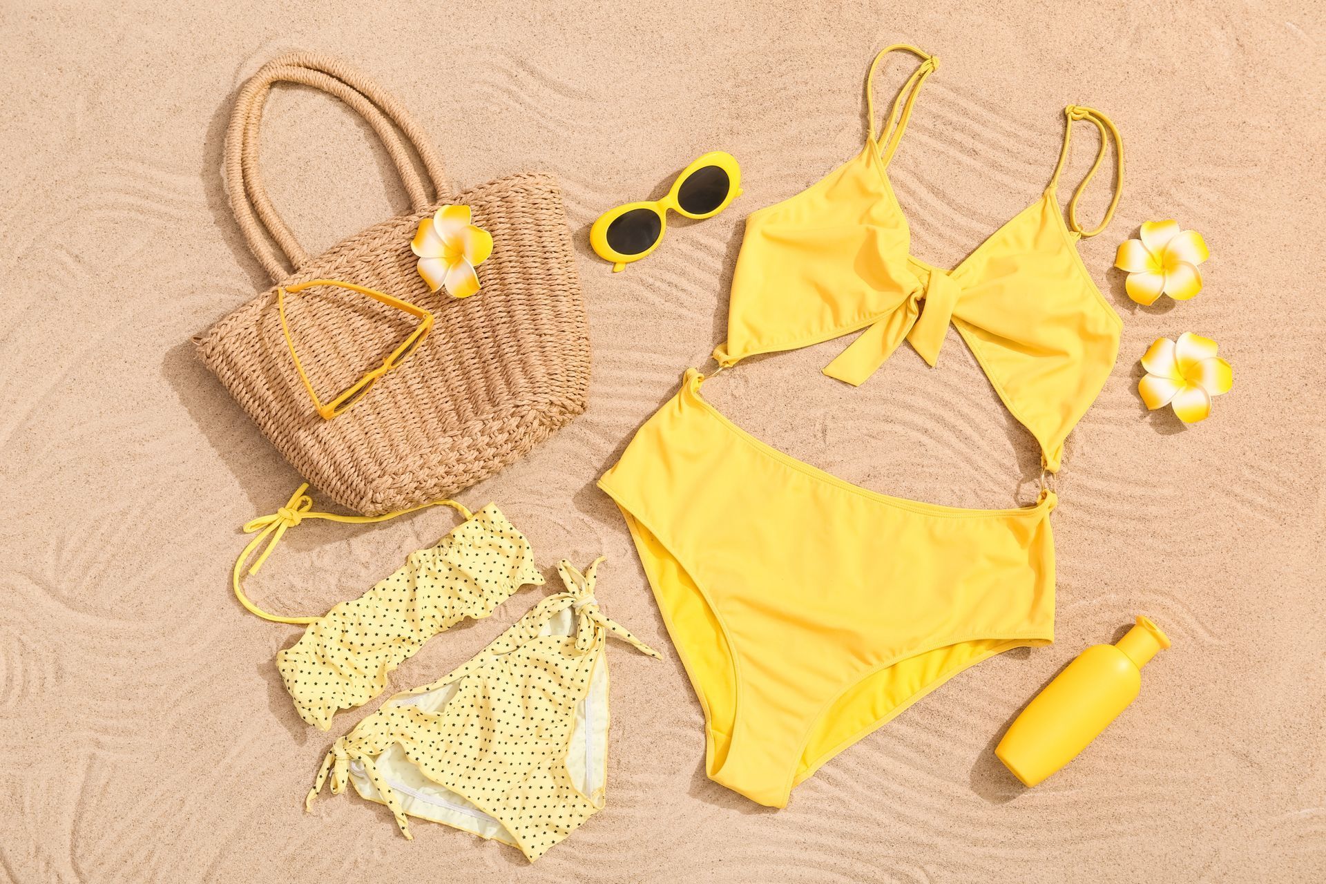 Yellow bikinis, straw beach bag, sunglasses, flower earrings, and sunscreen on a beige beach towel