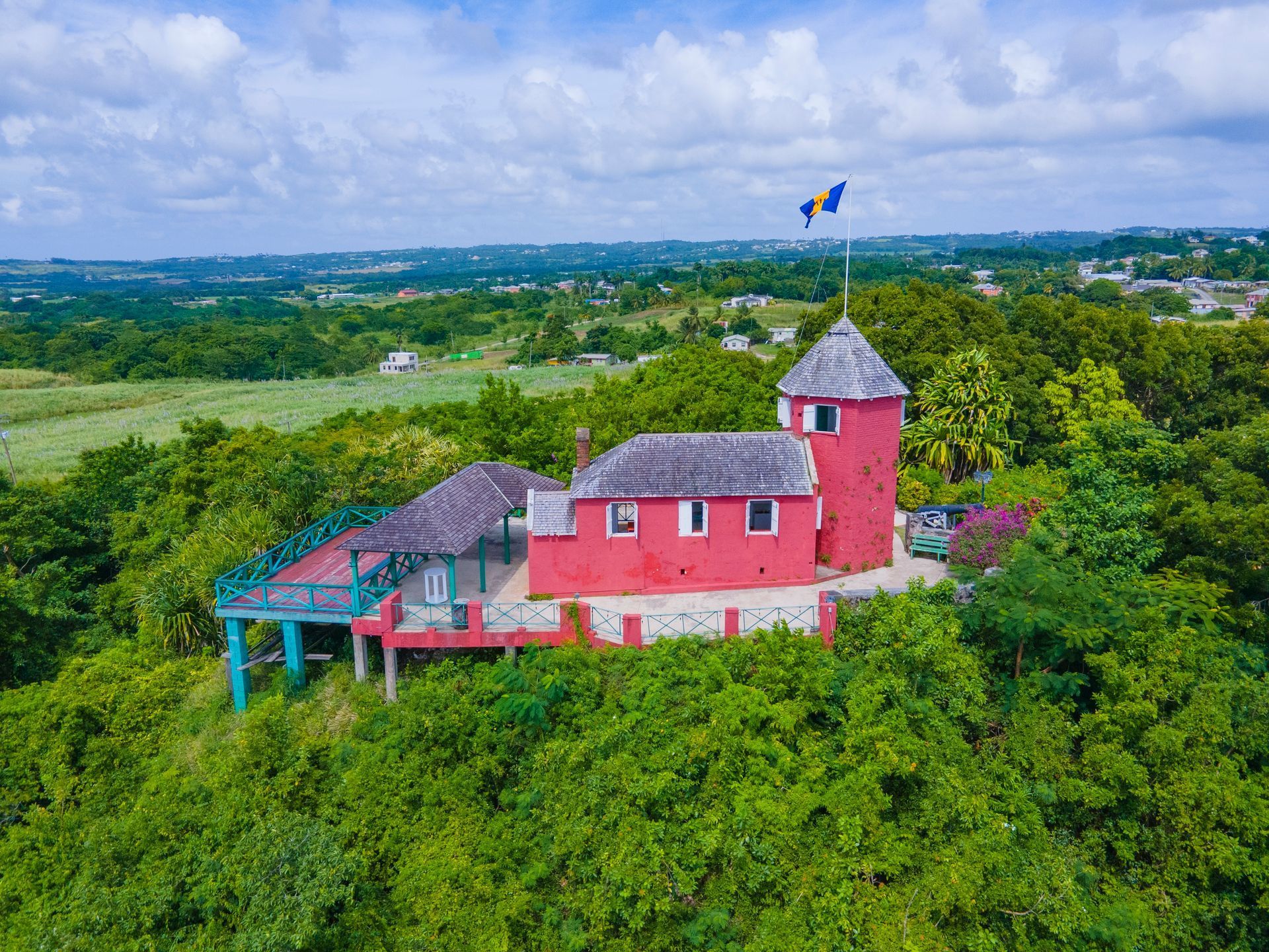 Red house with tower amid lush green hills under a blue sky, with a paraglider in the distance