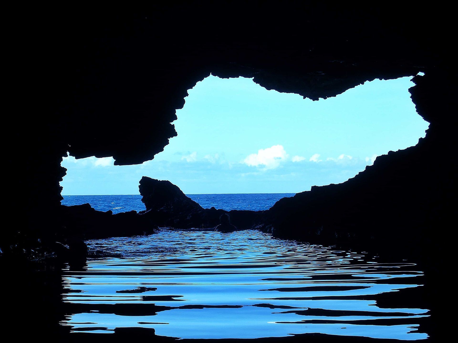 View from inside a dark cave toward bright blue sea and sky, with water rippling at the entrance
