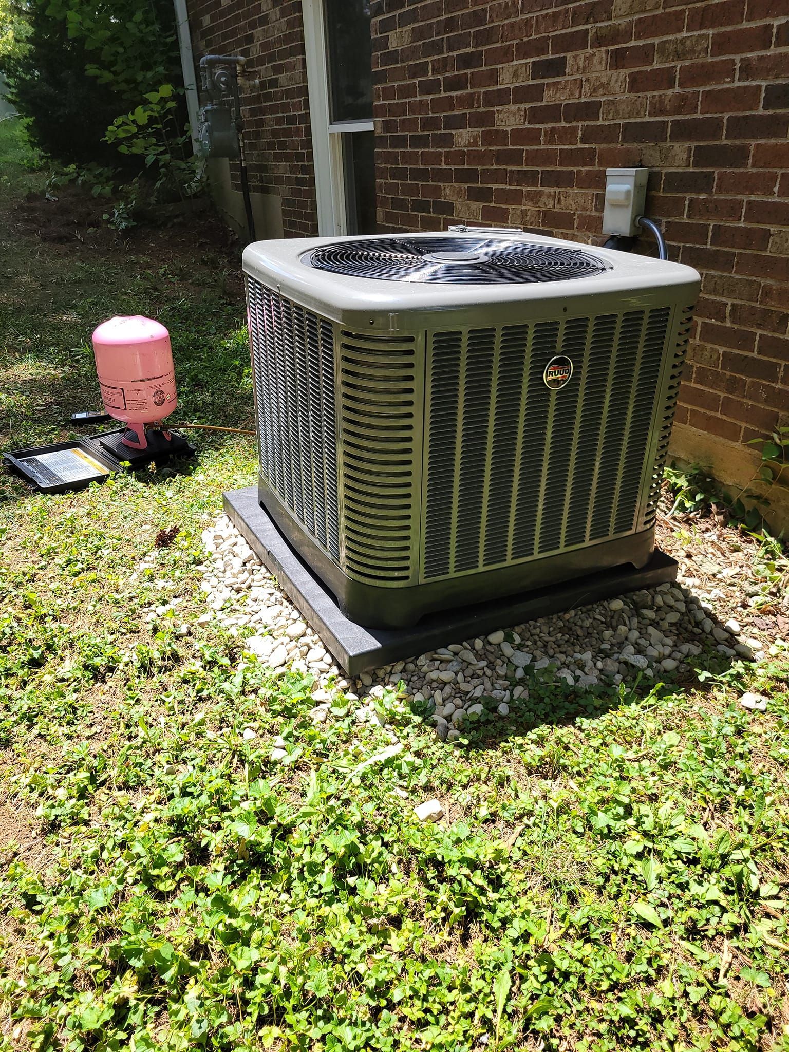 An air conditioner is sitting in the grass in front of a brick building.