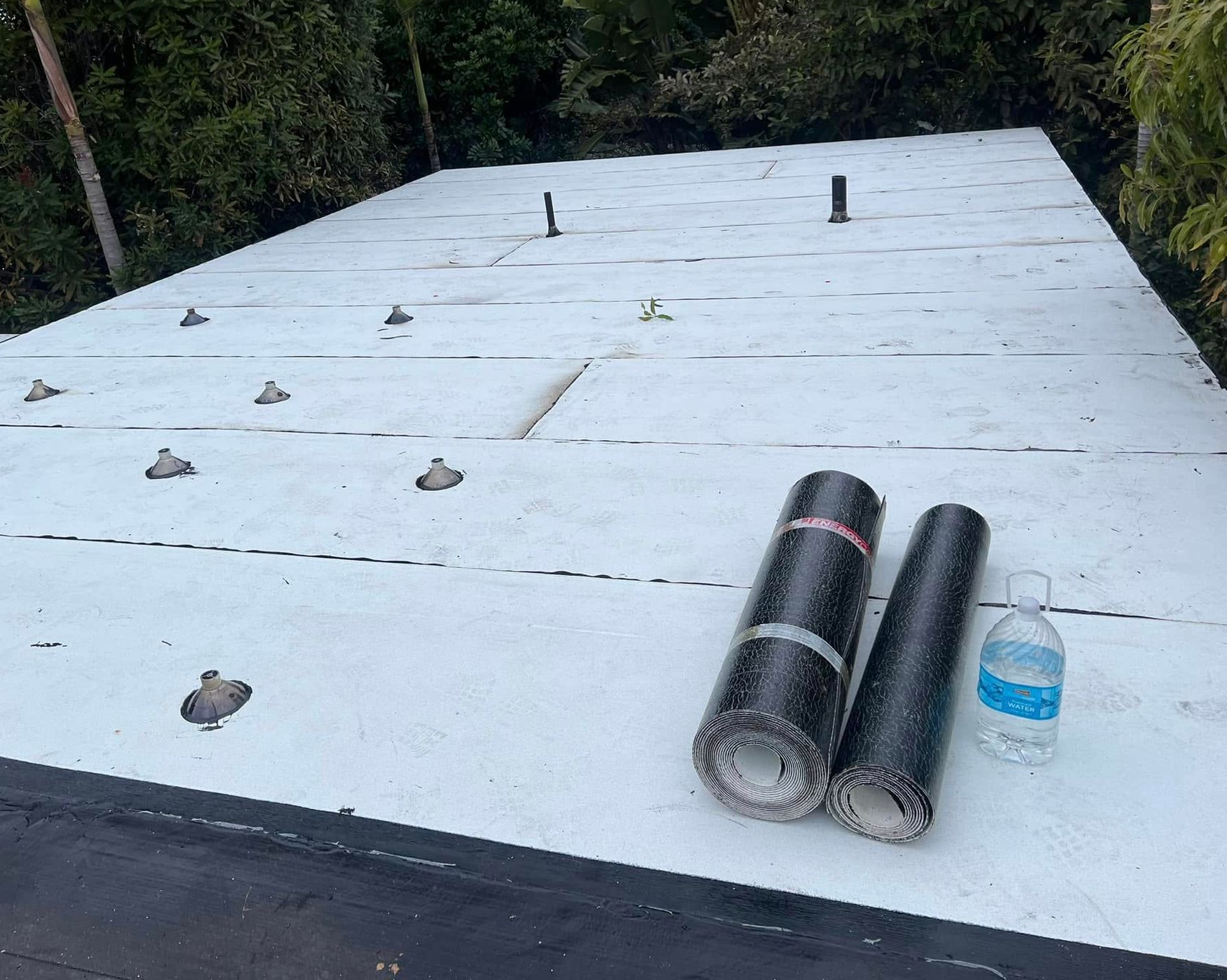 White rooftop under construction with scattered fasteners and two rolls of roofing material beside a water bottle.