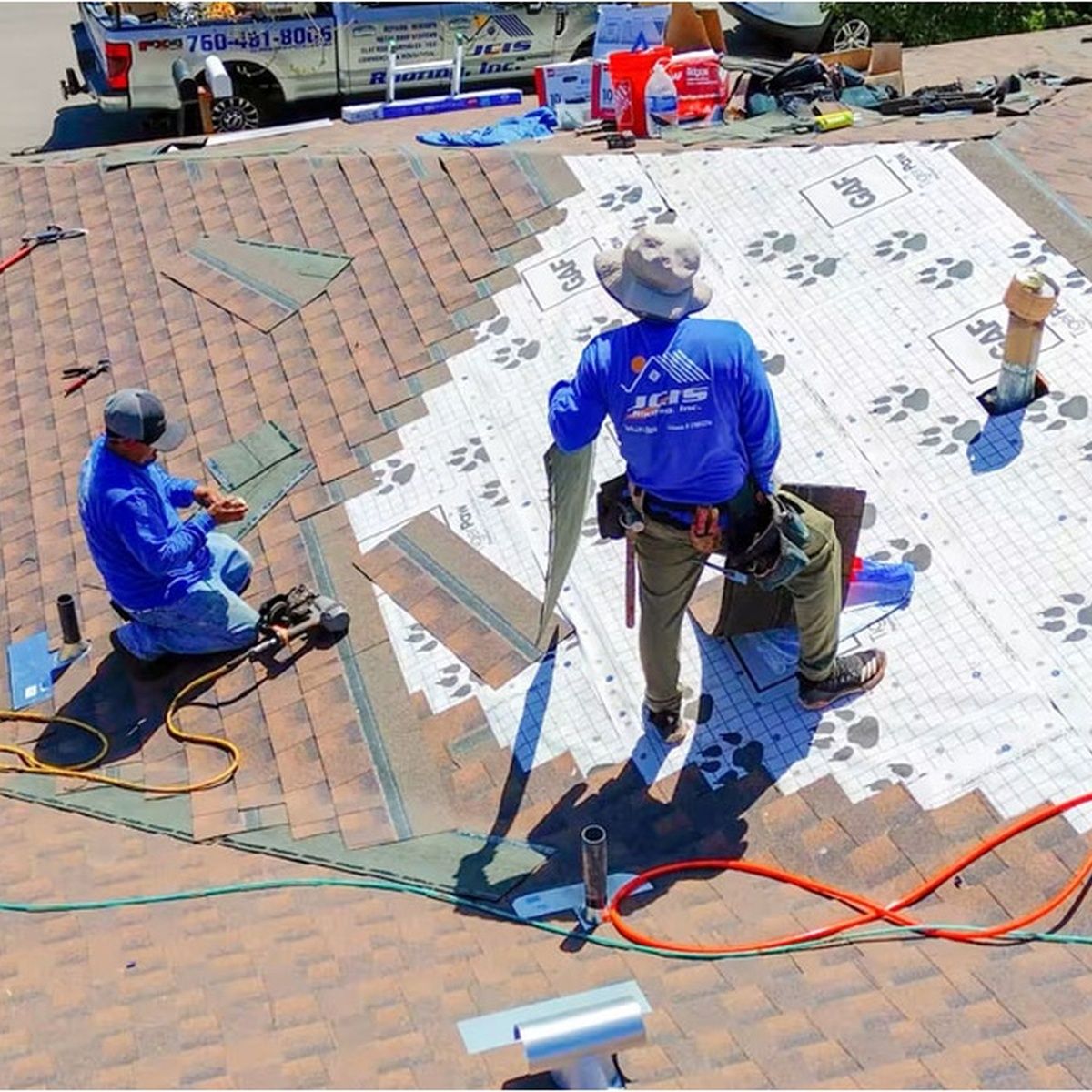 Workers repairing a shingled roof, with one standing and two kneeling beside tools and cords.