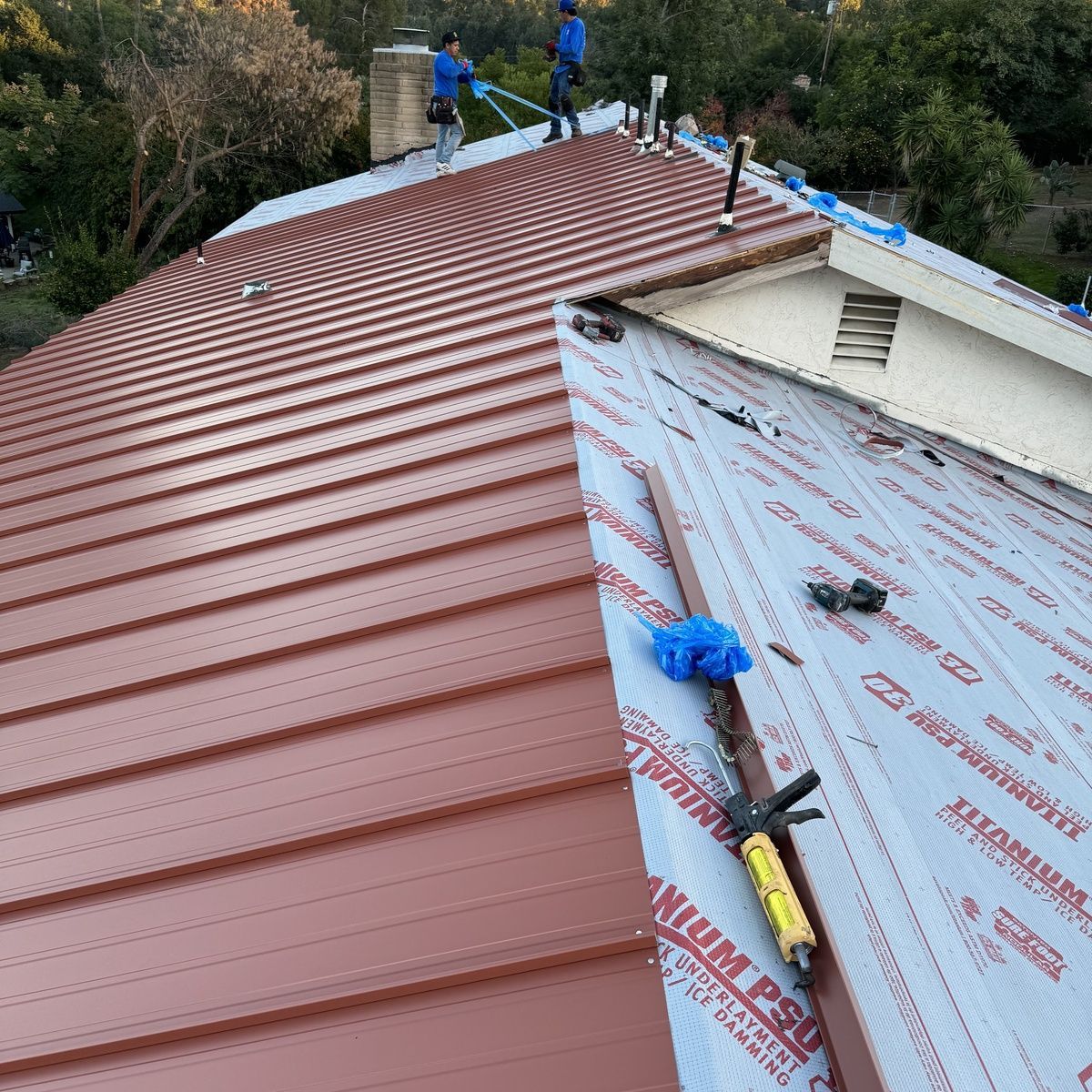 Workers installing red metal roofing on a steep roof with blue underlayment.