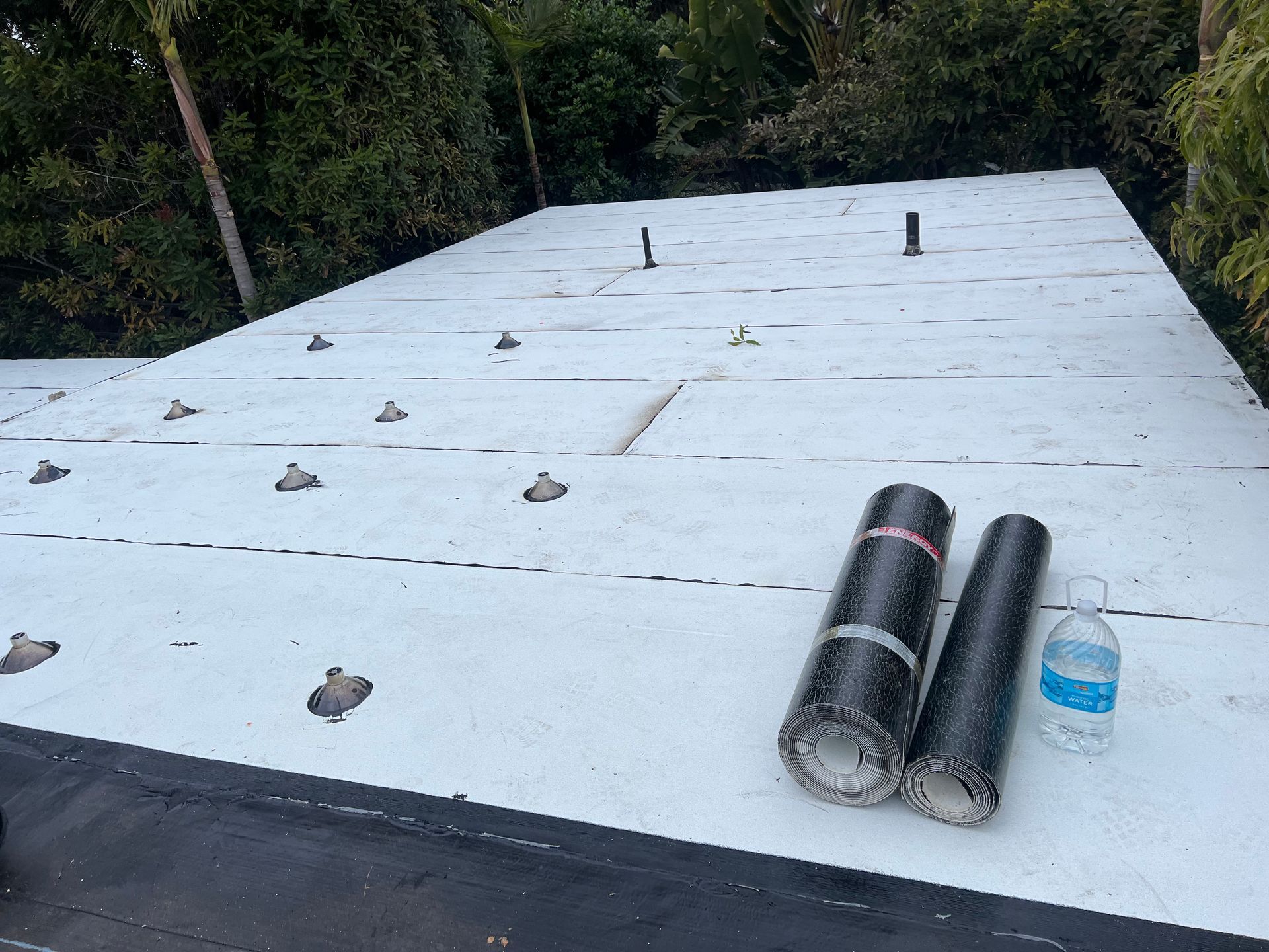 Flat white roof under construction with rolled roofing materials and trees in the background