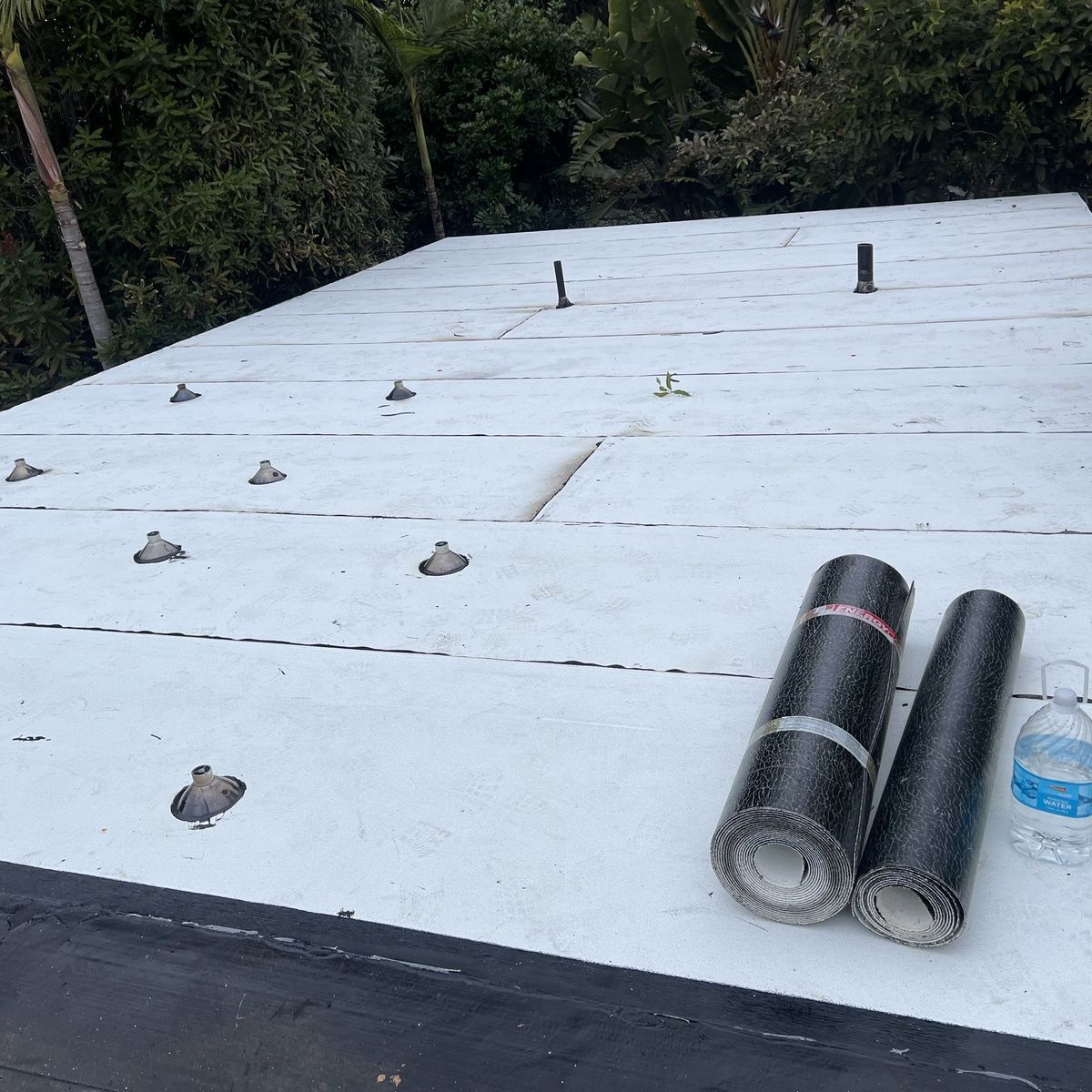White roof under construction with roofing membranes, vents, and a water bottle near a wooded area