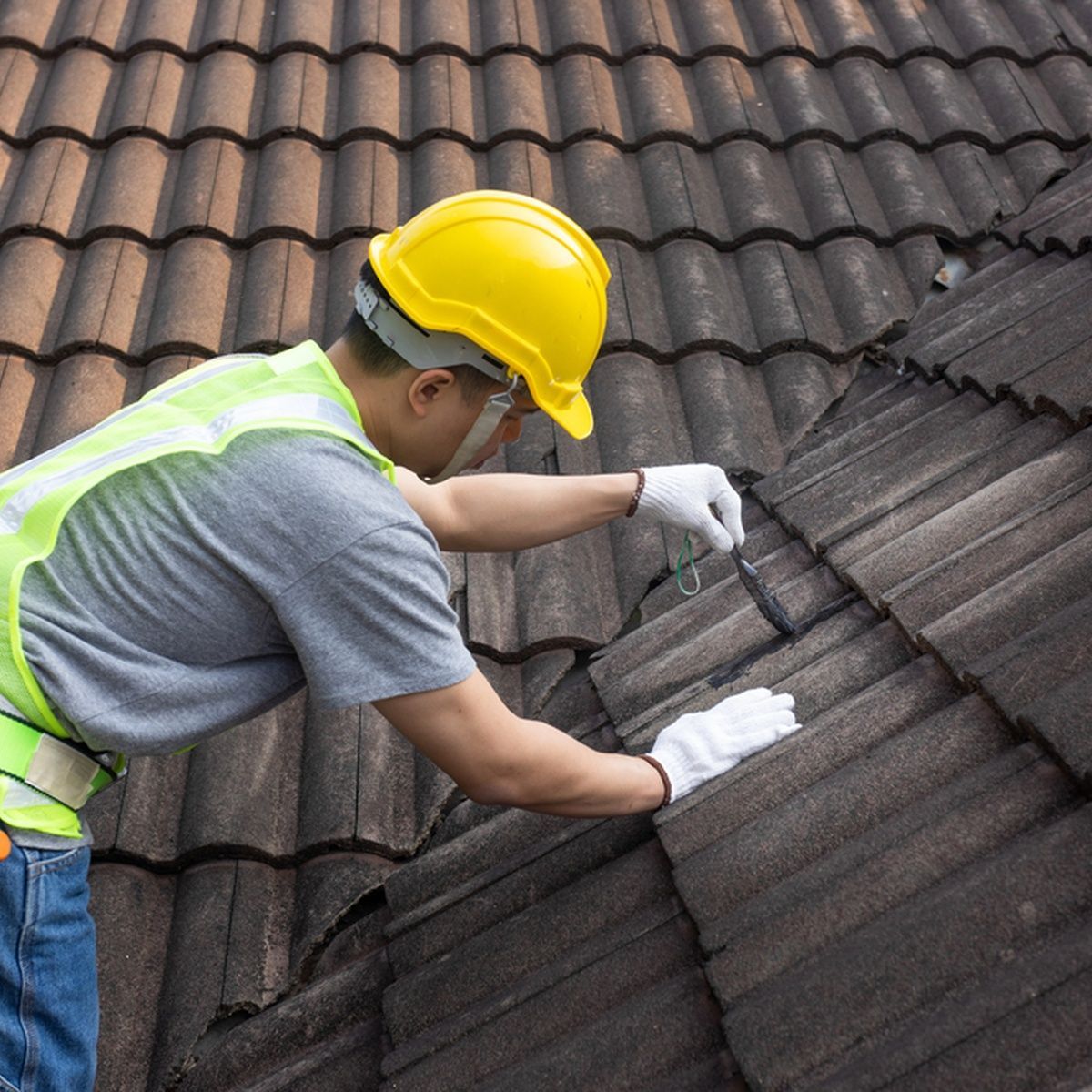 Worker in yellow hard hat repairing dark roof shingles