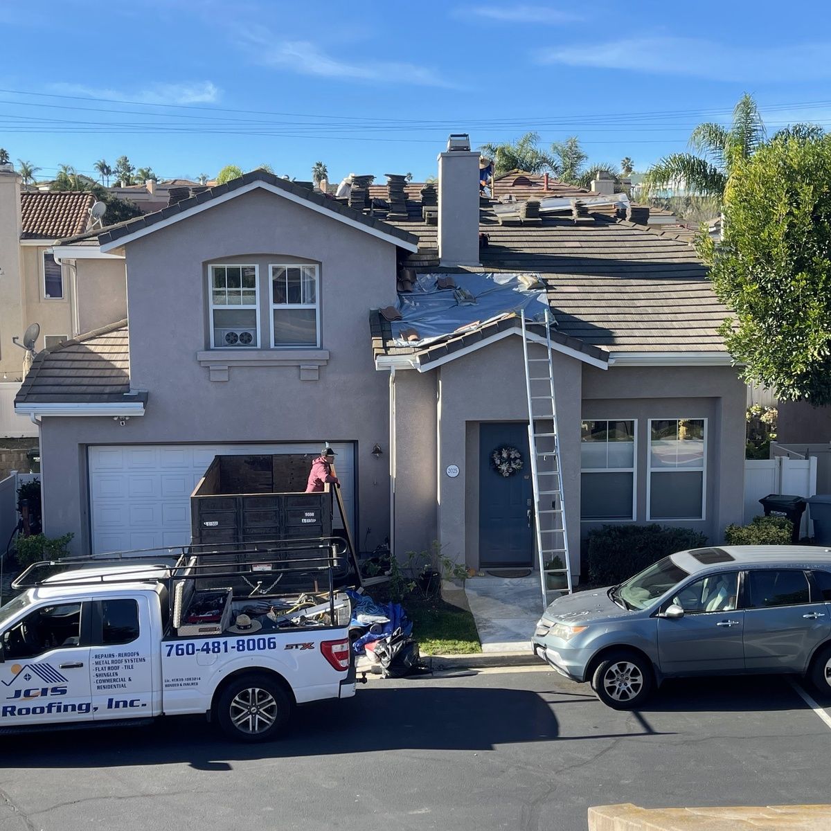 Two-story house with roof damage, ladders, and work trucks parked out front on a sunny street