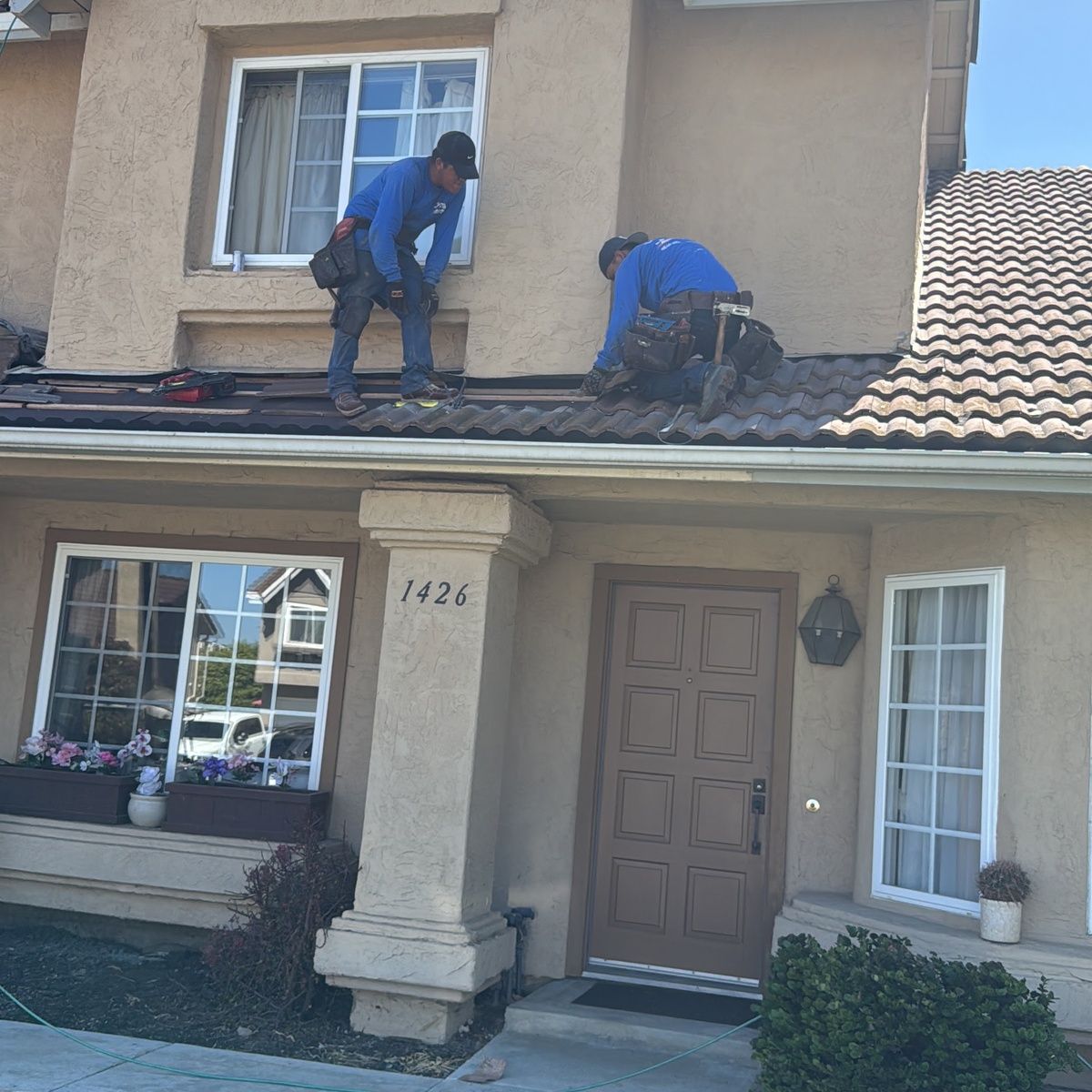 Two workers fixing roof tiles on a two-story house with a beige exterior and front porch.