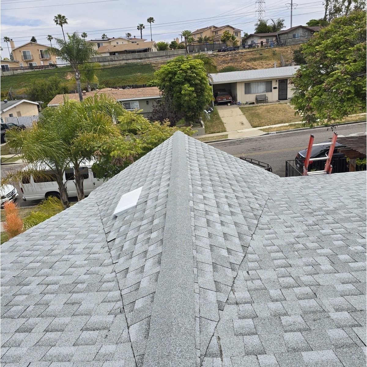 View from a shingled rooftop over a suburban neighborhood with houses, trees, and hills in the background