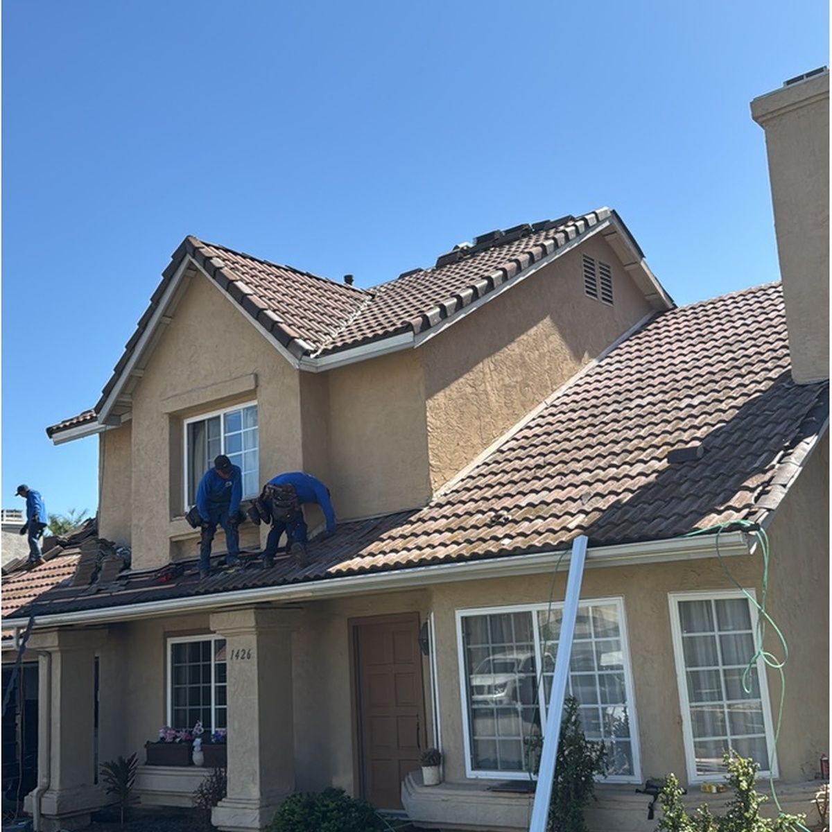 Two workers on a tan two-story house roof, repairing tiles under a clear blue sky