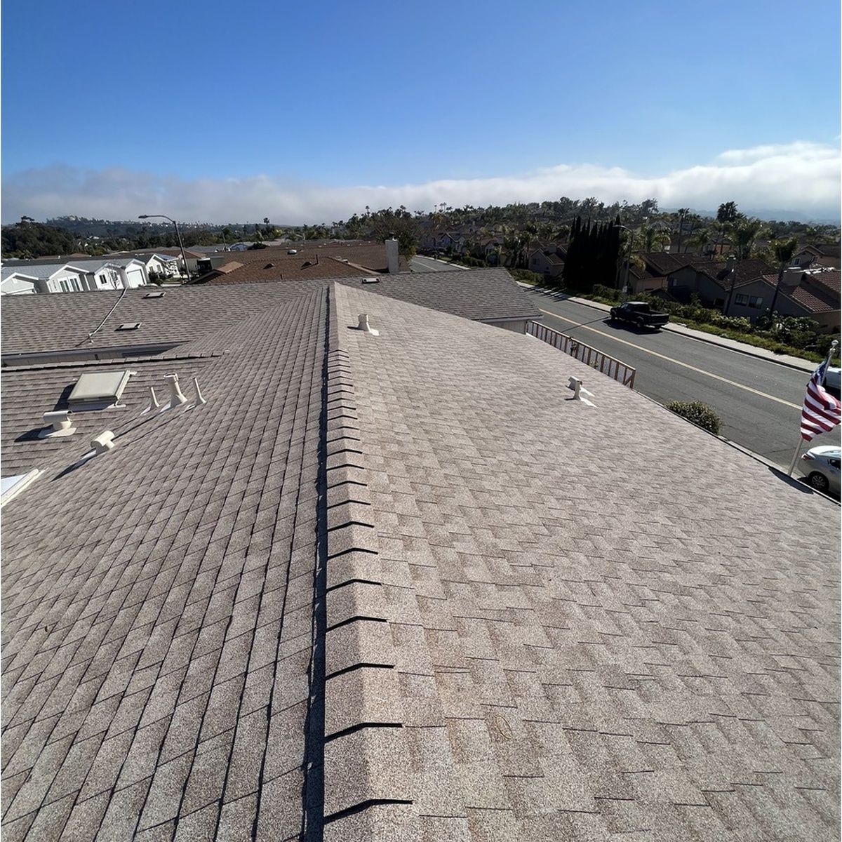 Rooftop shingles with a central ridge, overlooking a suburban neighborhood under a clear blue sky.