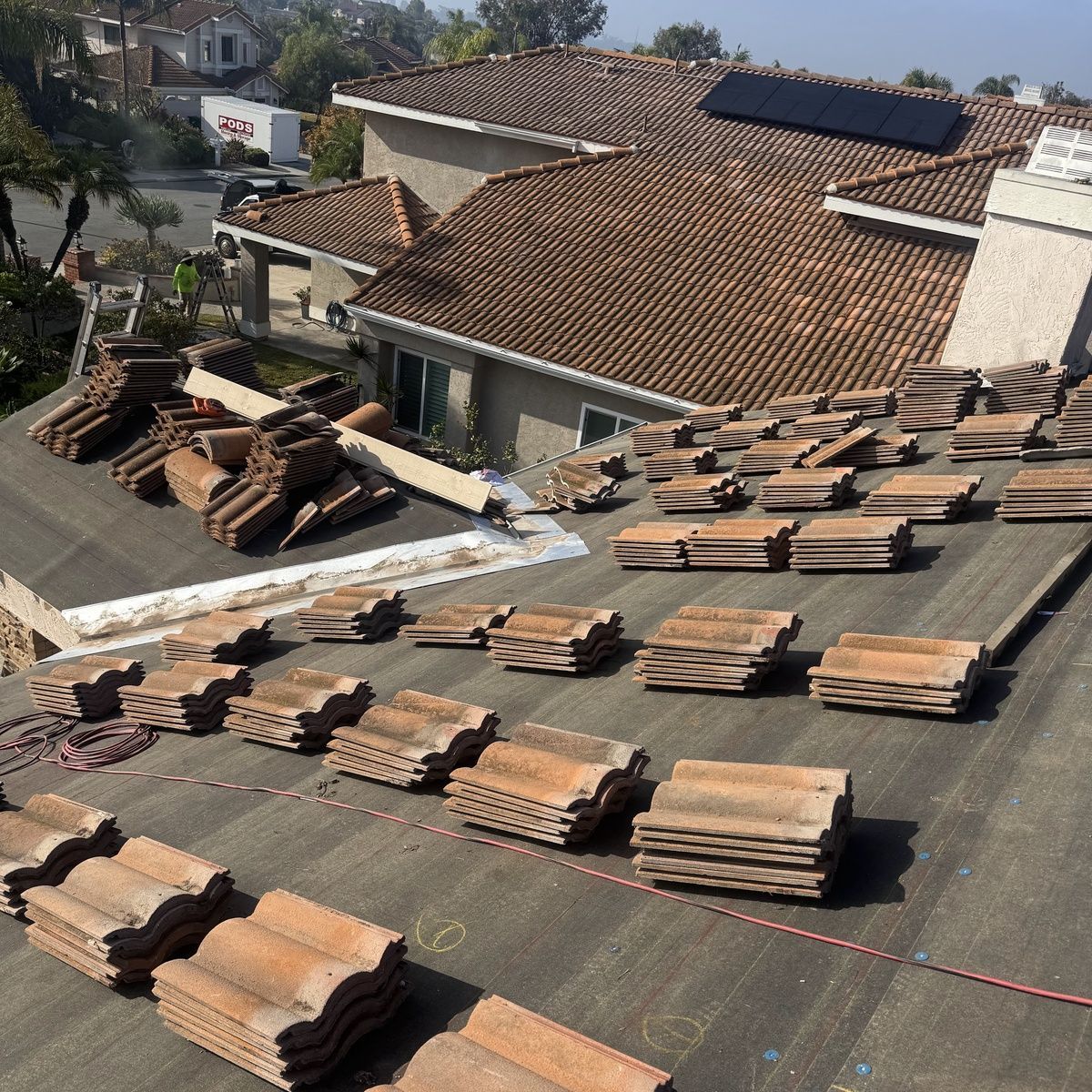 Rooftop covered with stacks of terracotta roof tiles under construction, with houses in the background.