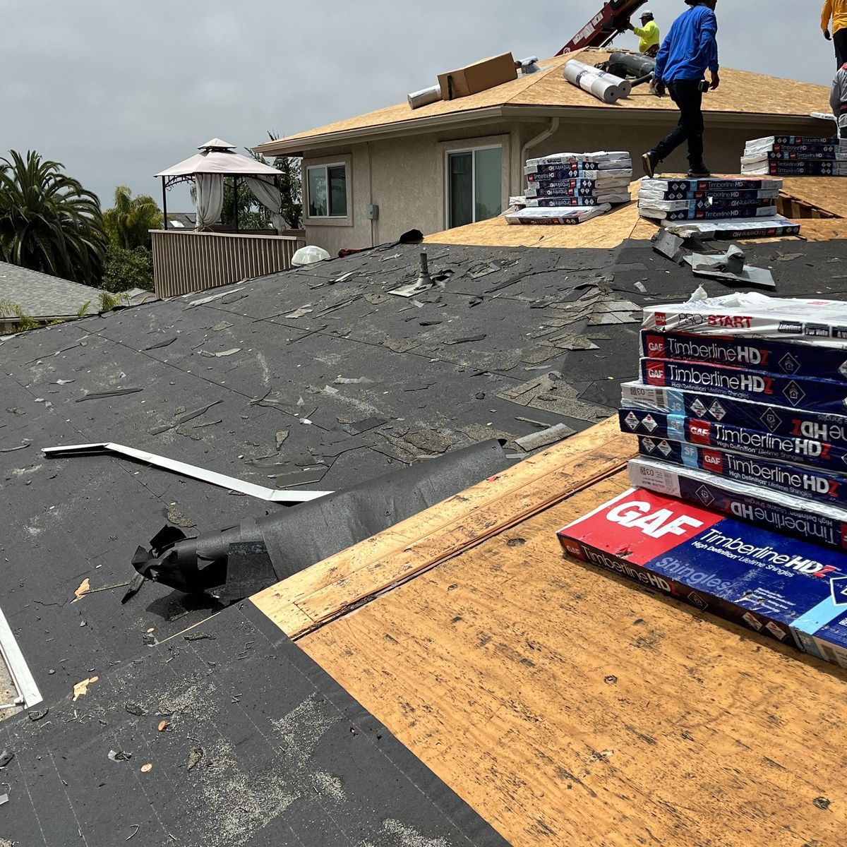 Roofers installing shingles on a house roof, with stacks of roofing materials and plywood visible.