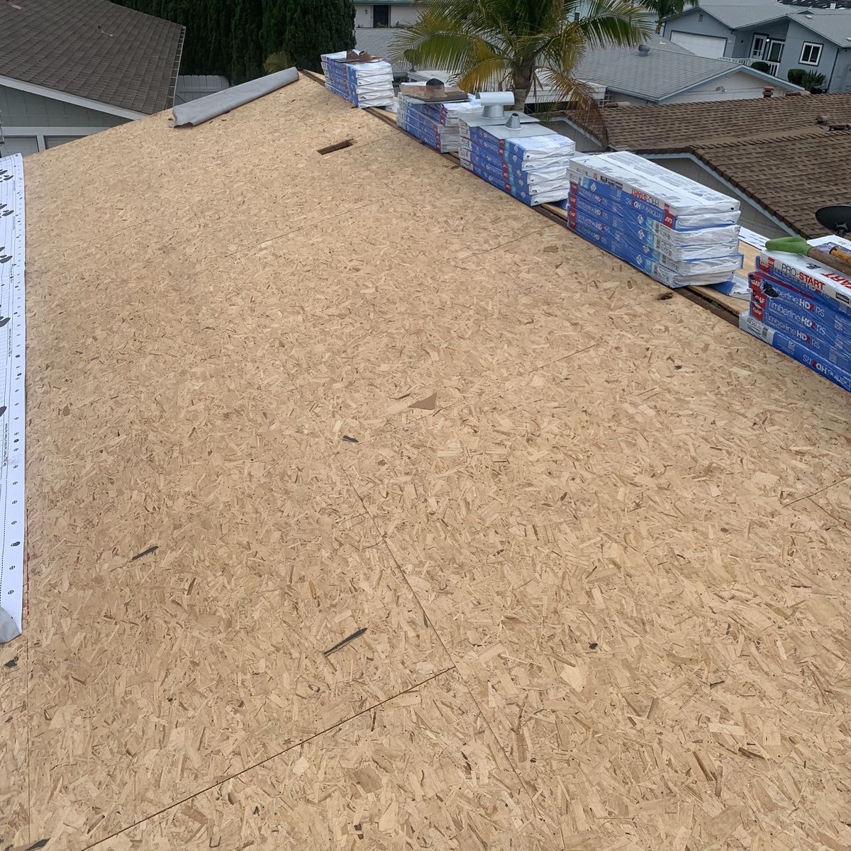 Roof under construction with plywood sheathing and stacks of roofing materials along the edge