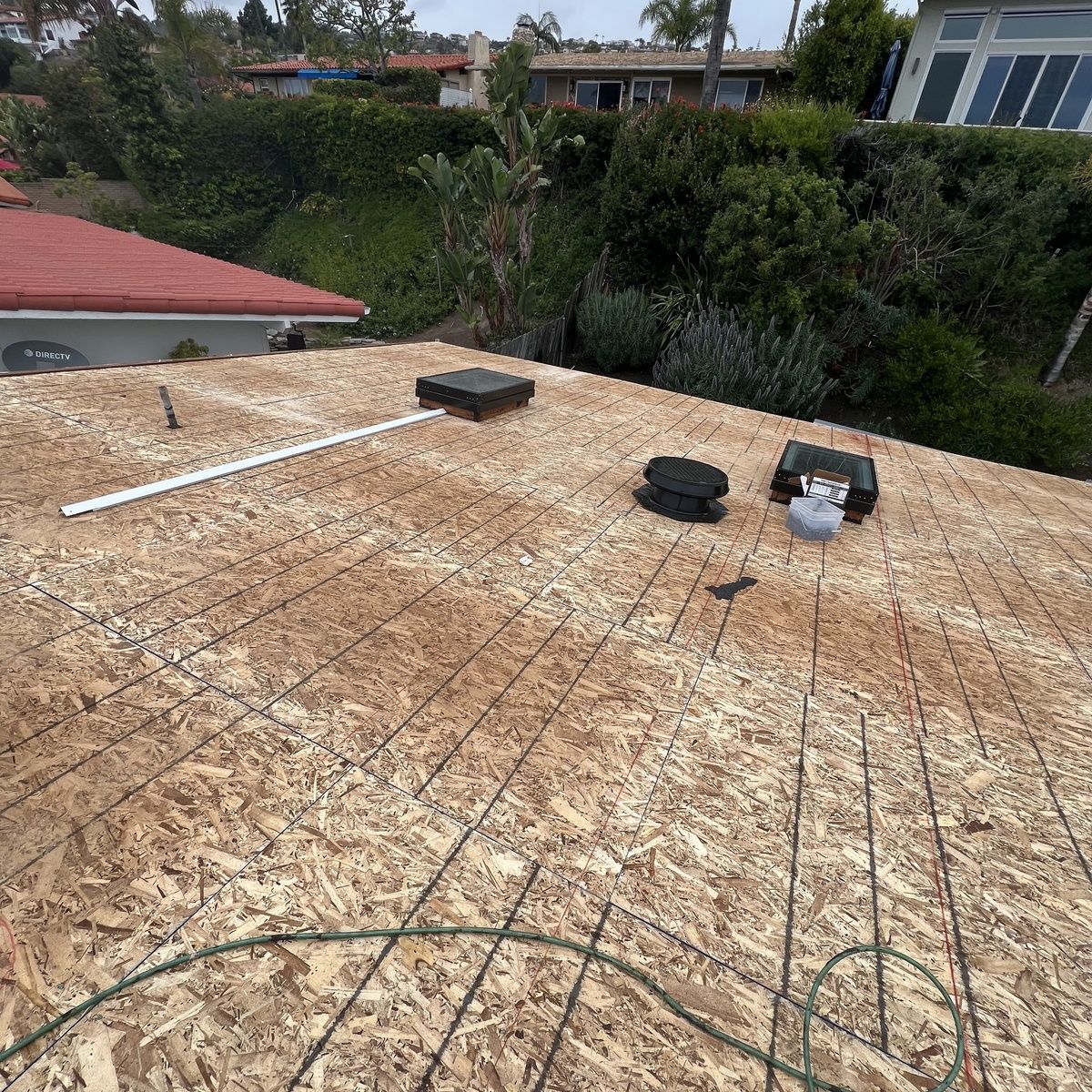 Rooftop under construction with wood sheathing, rebar, and a few tools, overlooking trees and houses.