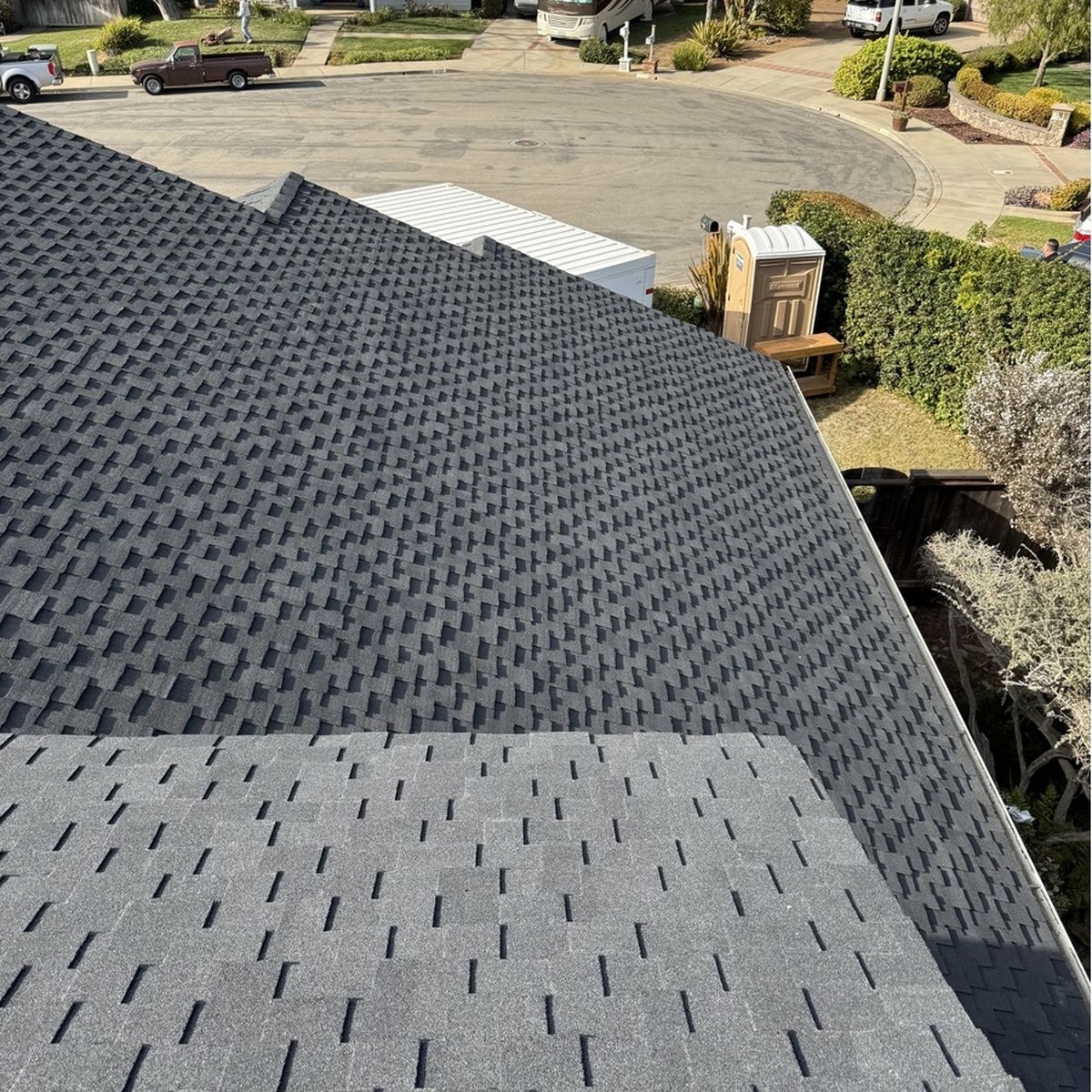 Dark gray roof shingles with part of a lighter patch, overlooking a suburban driveway and yard below