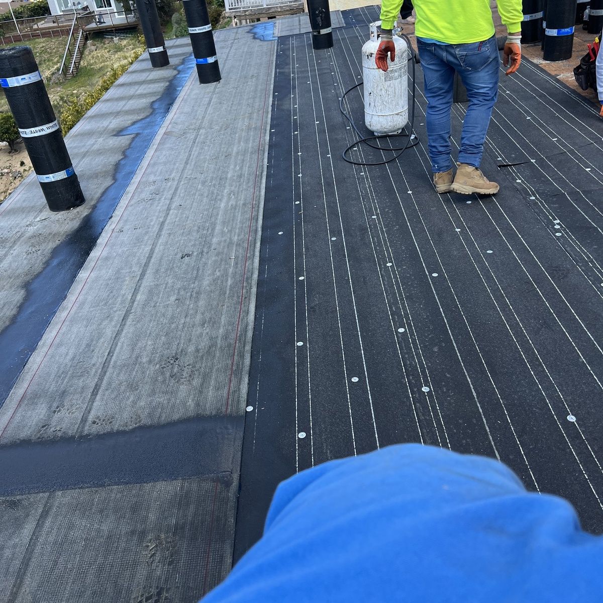 Workers laying black roofing membrane on a flat roof with white guide lines and black vents.
