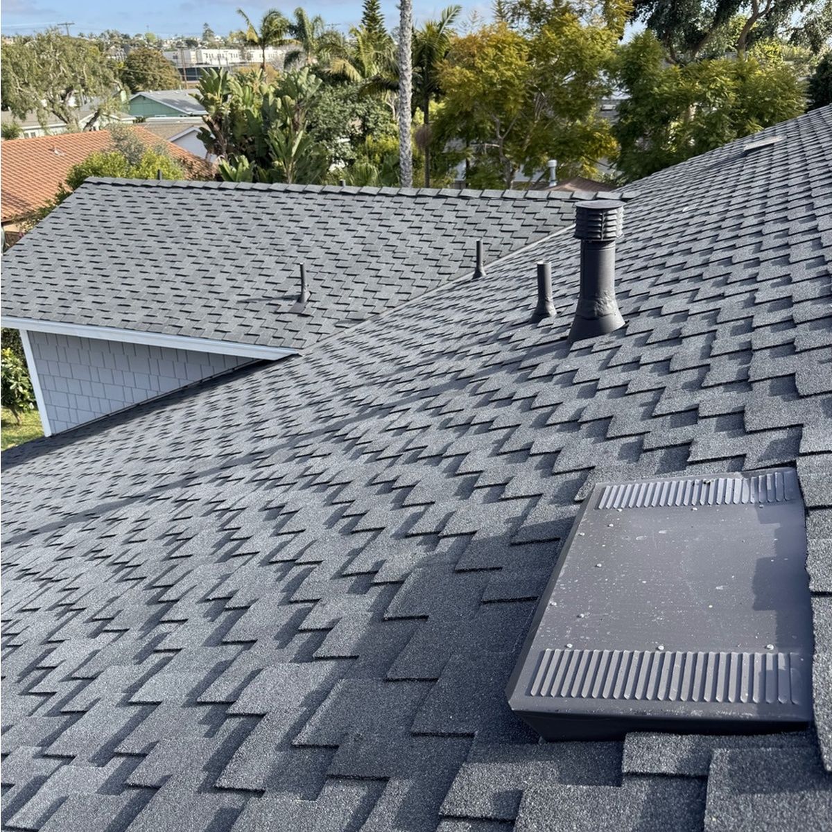 Rooftop with dark gray shingles, vent pipes, and a skylight, viewed from above with trees in the background