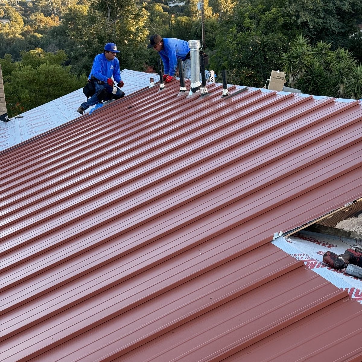 Workers installing a red corrugated metal roof on a house, with trees in the background.