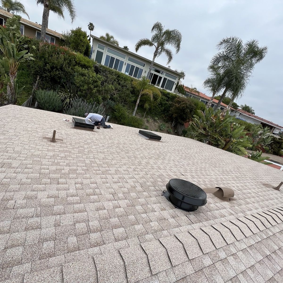 Worker on a tiled rooftop among vents, with houses, trees, and a cloudy sky in the background