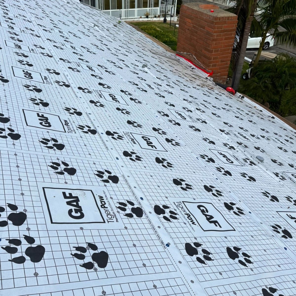 Roof covered with white paw-print underlayment, with a brick chimney and house edge visible.