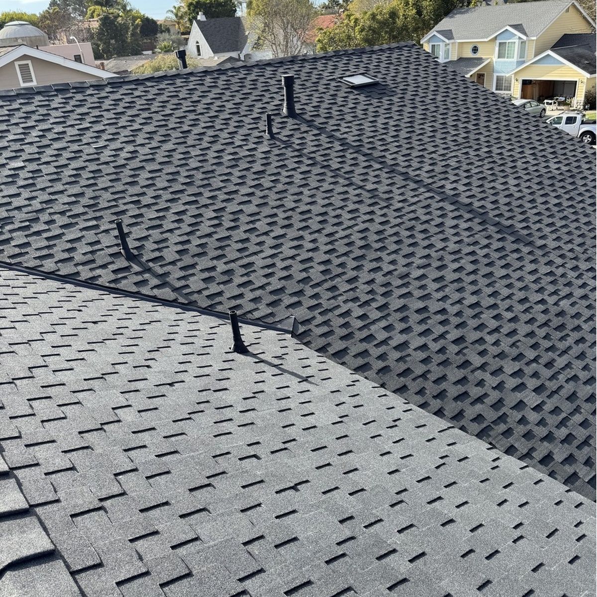 Two shingled roof sections on a house, with vent pipes and neighboring homes in the background