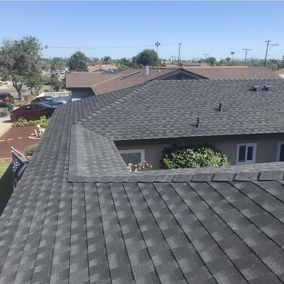 Black shingle roof of a suburban house under a bright sky, with neighboring homes and trees nearby