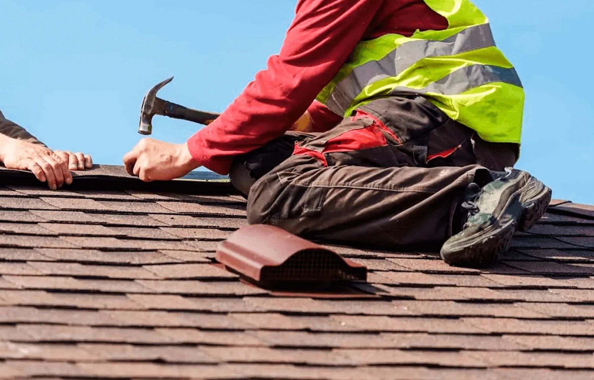 Roofing worker in red shirt and reflective vest nailing shingles on a rooftop
