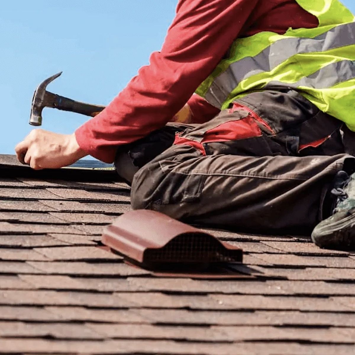 Roofer in red shirt and safety vest hammering shingles on a roof