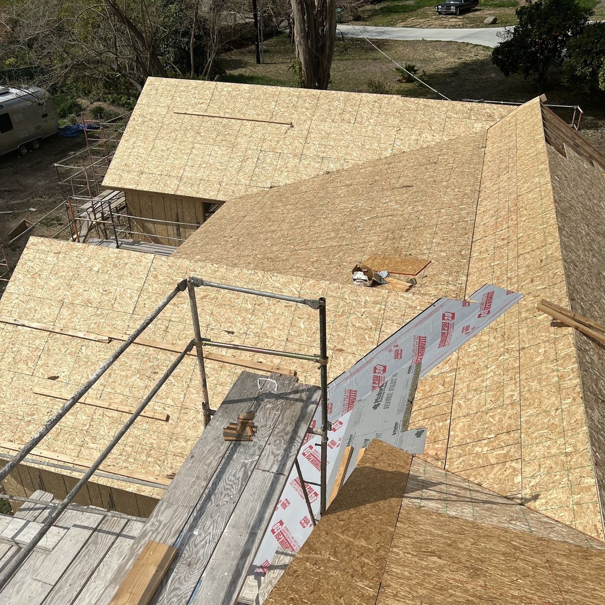 Roof under construction with plywood sheathing, scaffolding, and wood framing on a house in a wooded area