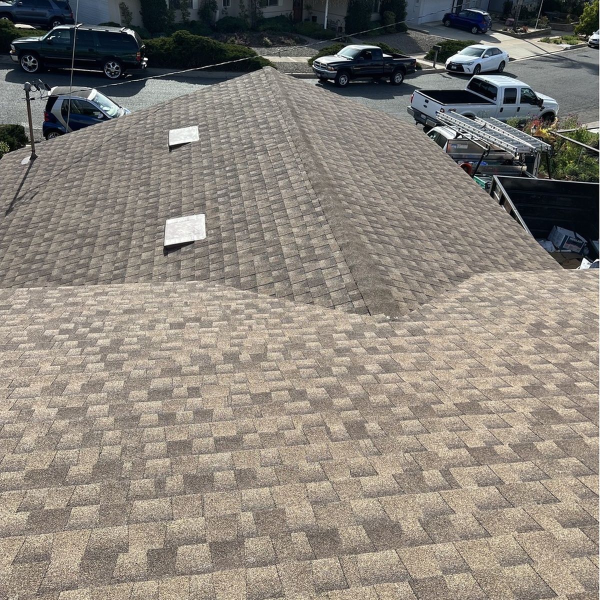 Rooftop covered in brown shingles with skylights, viewed from above near parked cars and houses.