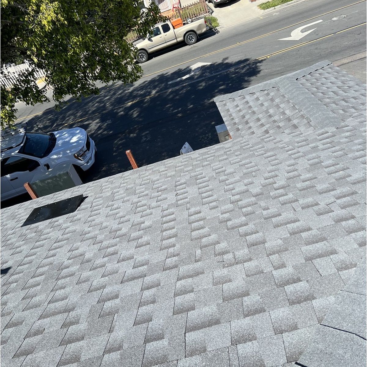 Steep gray shingled rooftop overlooking a tree-lined street with parked cars and a white truck