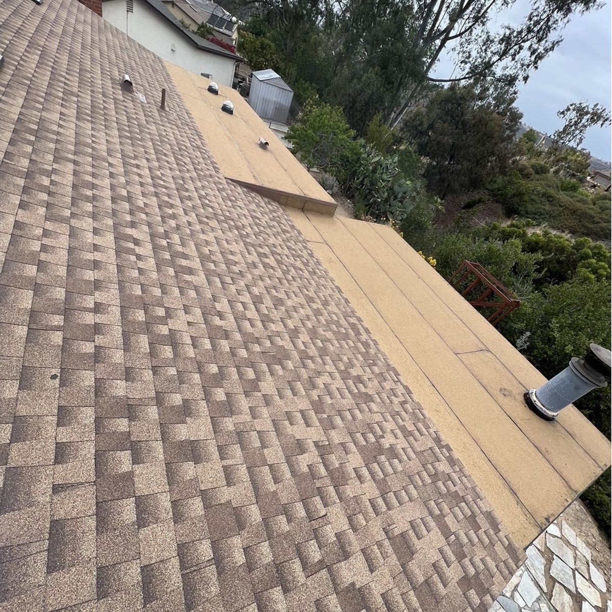 Tilted view of a shingled roof with rooftop vents, trees, and a cloudy sky beyond