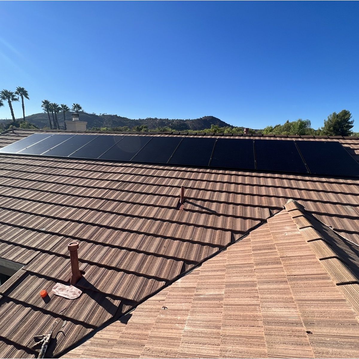 Solar panels on a tiled rooftop under a clear blue sky, with mountains and palm trees in the distance.
