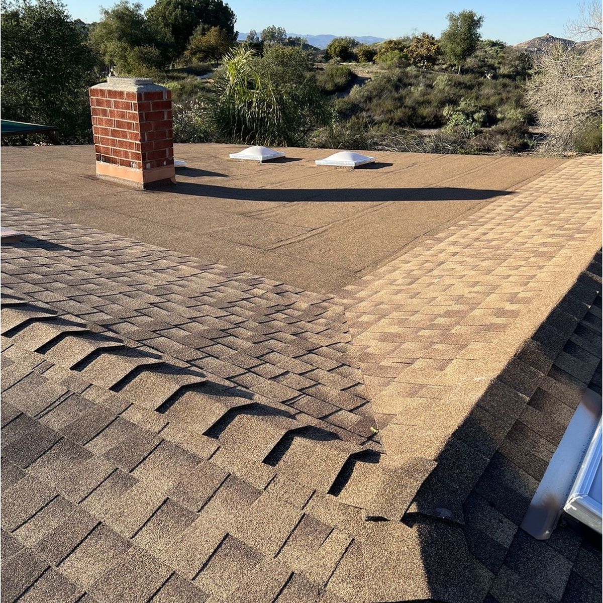 Flat roof with shingles, a red brick chimney, and two white vents against a wooded background.