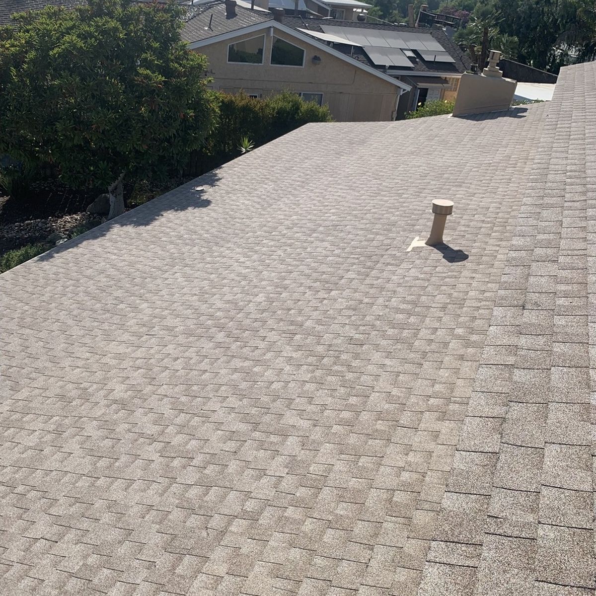 Sunlit gray shingle roof with a vent pipe, viewed from above with houses in the background