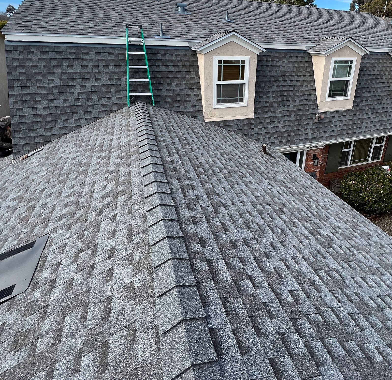 Roof with gray shingles and a green ladder leaning to the upper roof near dormer windows