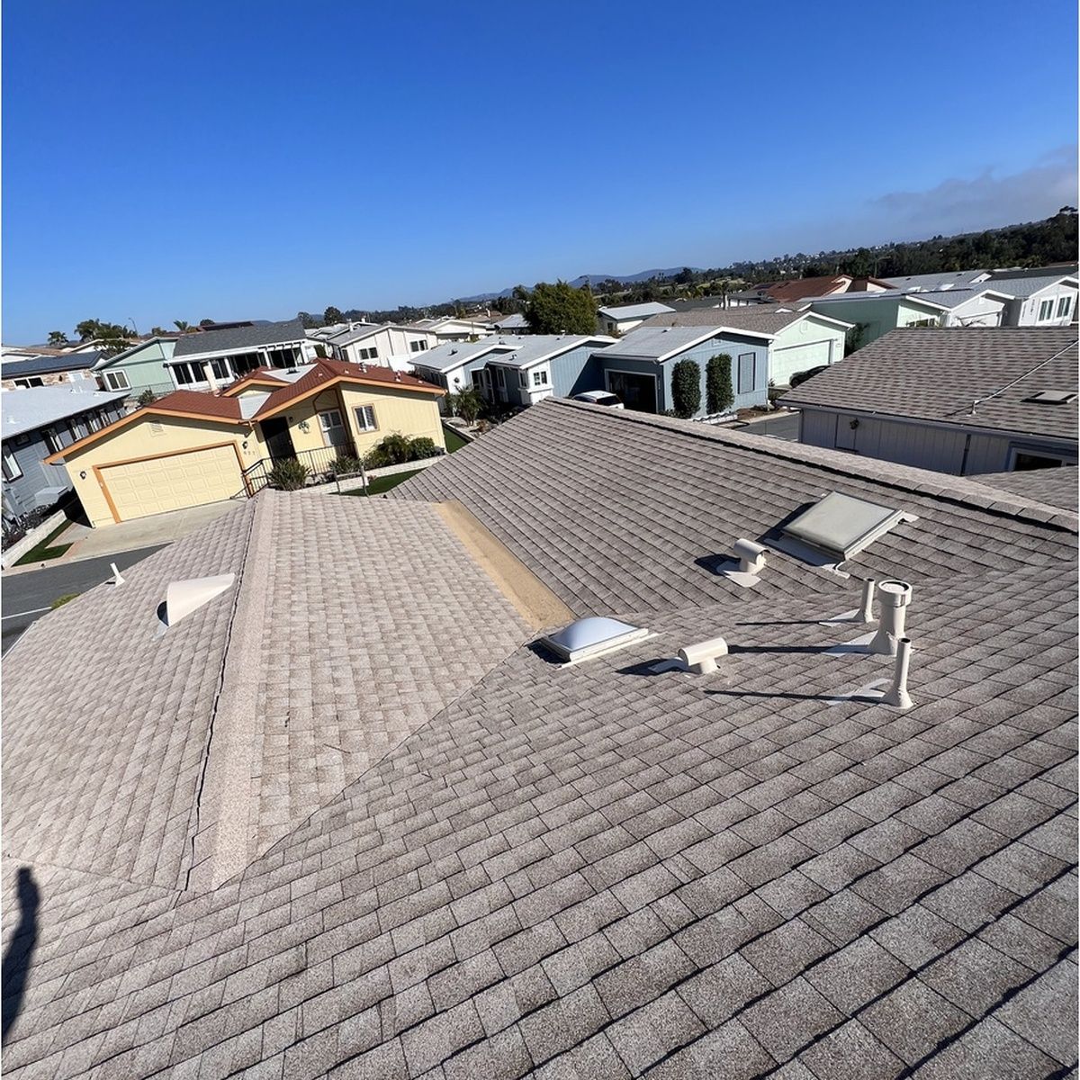 View across a suburban neighborhood from a shingled rooftop under a clear blue sky
