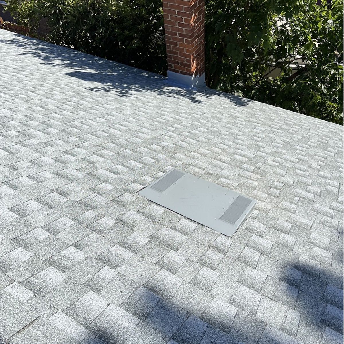 Gray shingled roof with a small access panel near a brick chimney and trees in the background