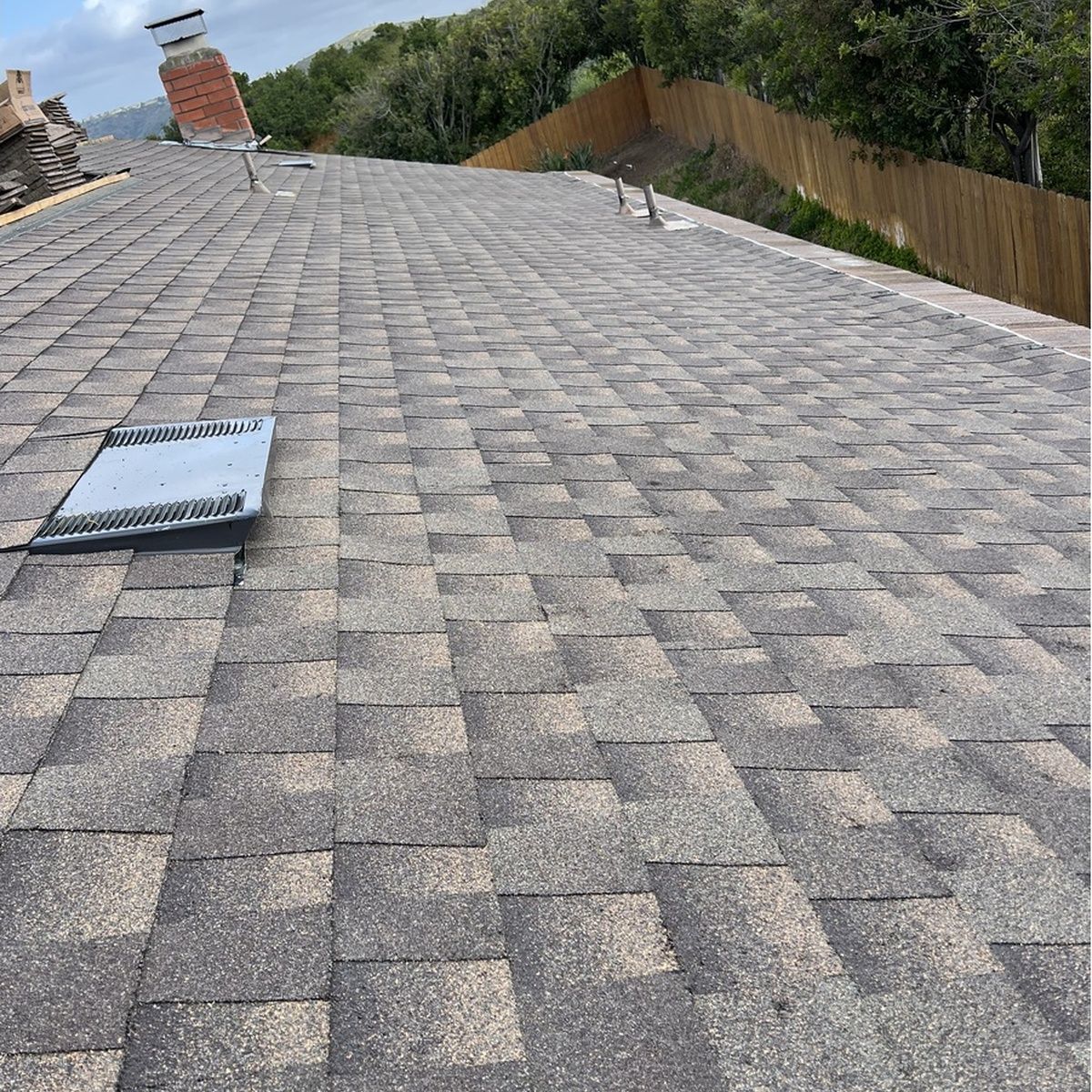 Roof covered in gray shingles with a vent opening, sloping down toward trees and a distant building
