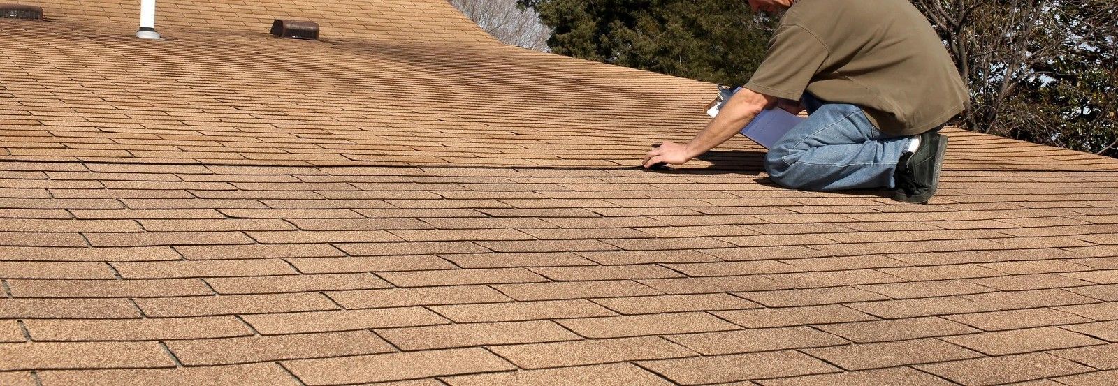 Gray shingle roof with vents, viewed from above, with houses, trees, and cloudy sky in the background