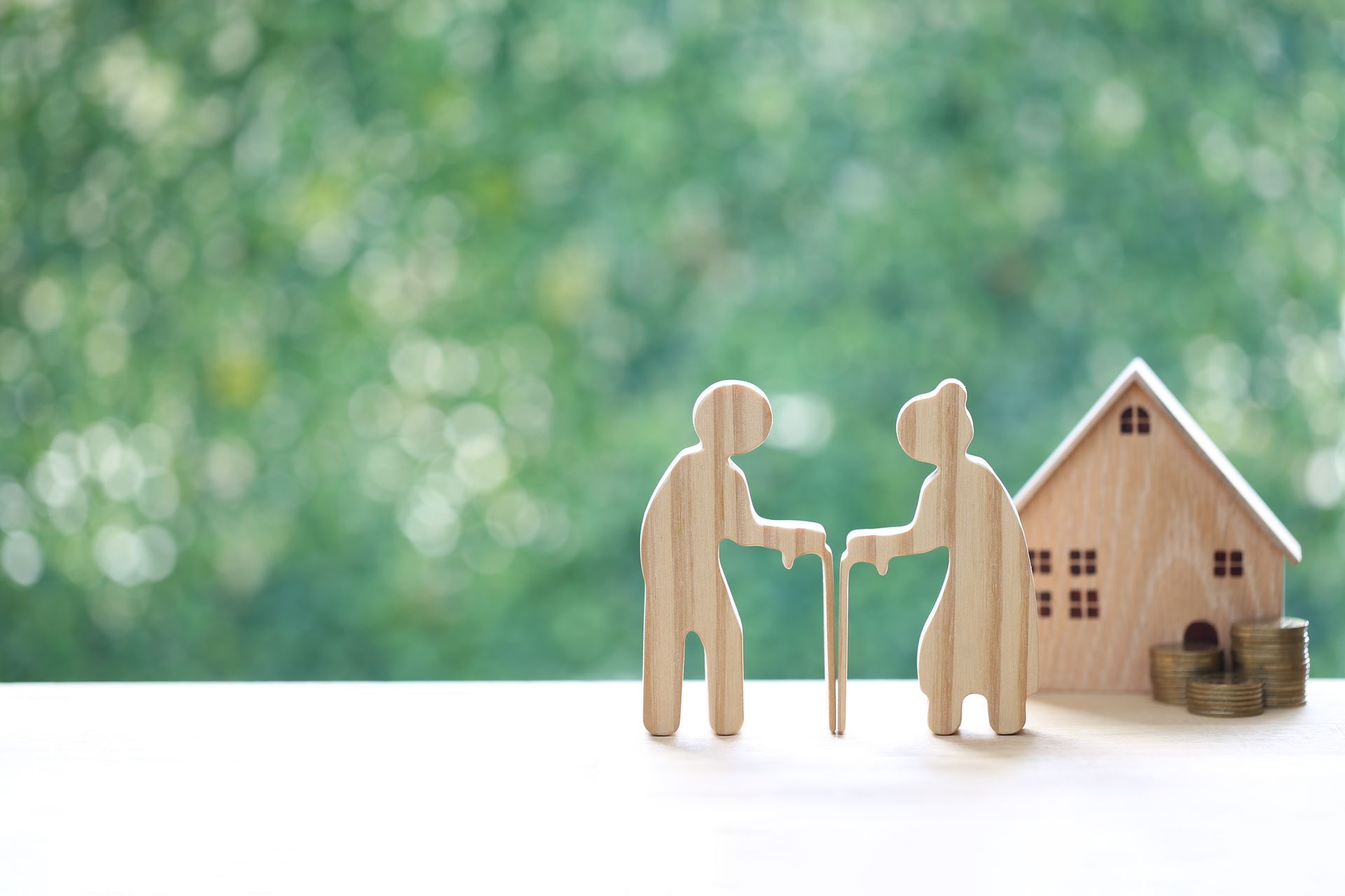 Two wooden figures shaking hands beside a small house and stacked coins on a white surface, green blurred background
