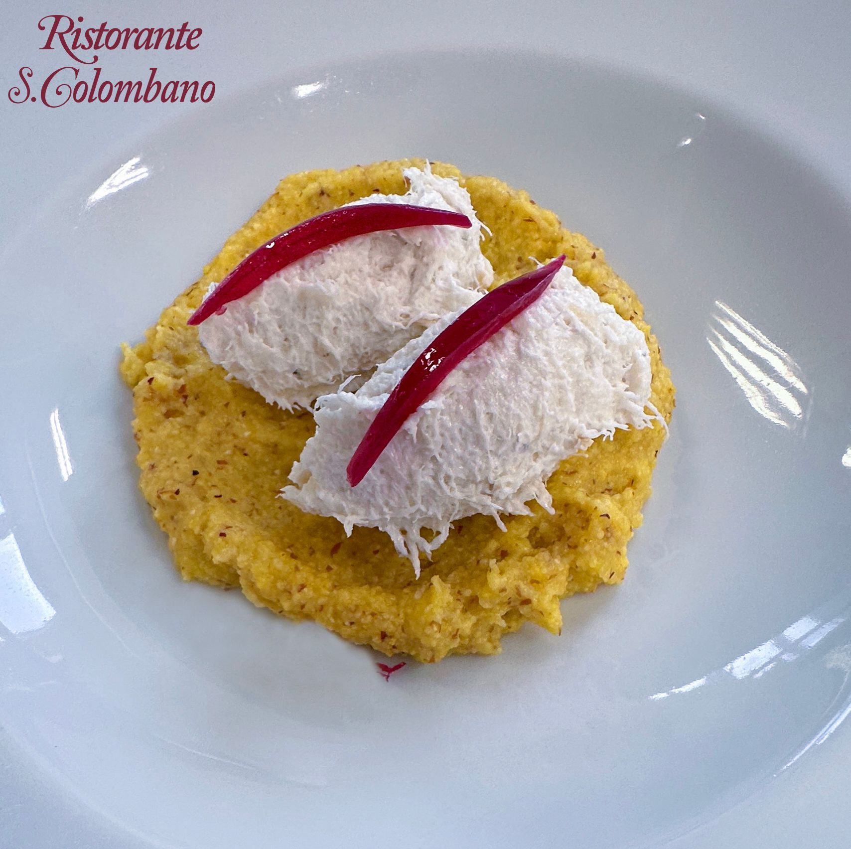 Due quenelle di pesce bianco guarnite con foglie di bietola rossa, servite su un letto di polenta gialla in una ciotola bianca.