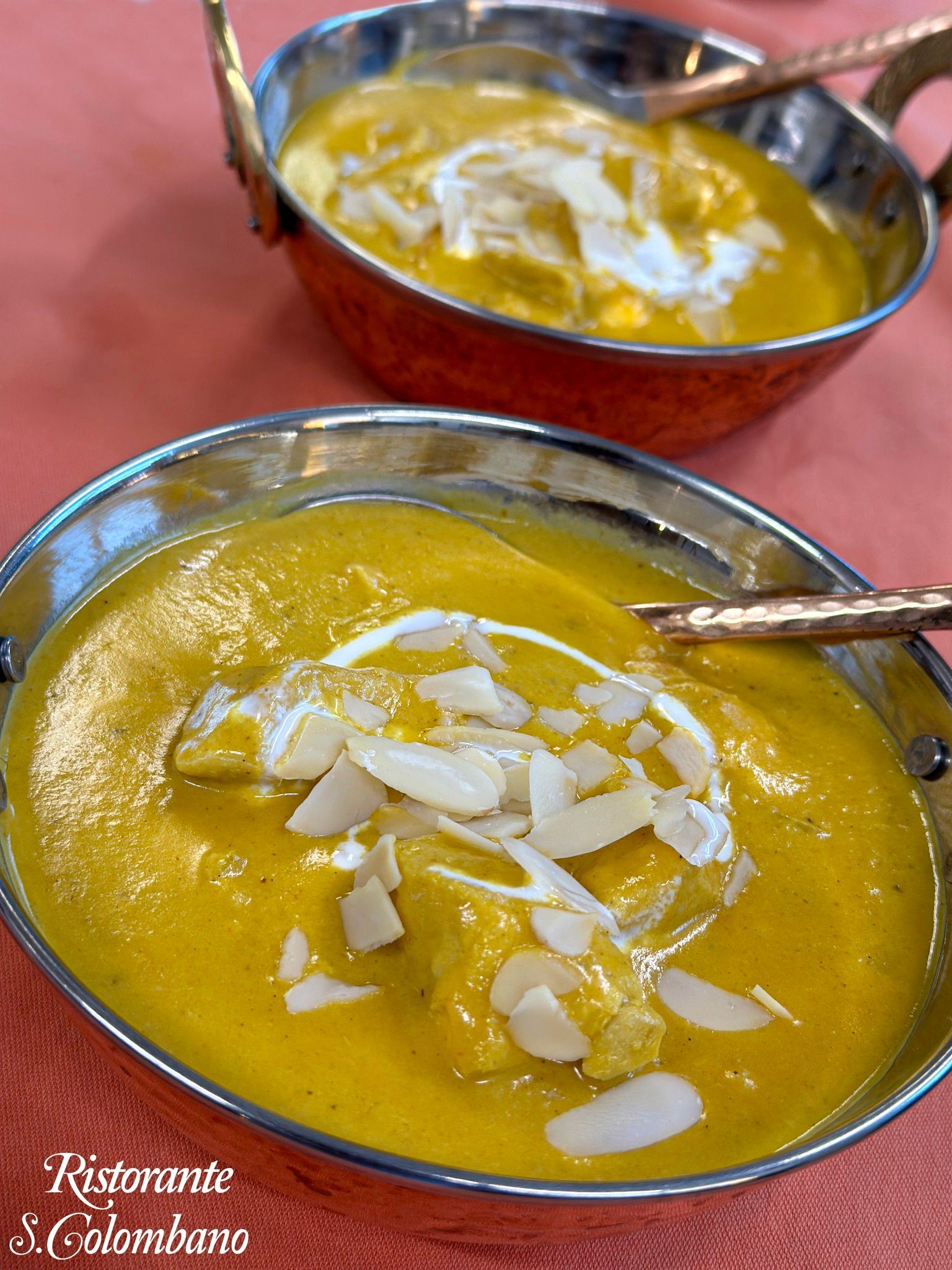 Due ciotole di curry indiano giallo guarnite con panna e mandorle a lamelle, servite su una tovaglia arancione chiaro.