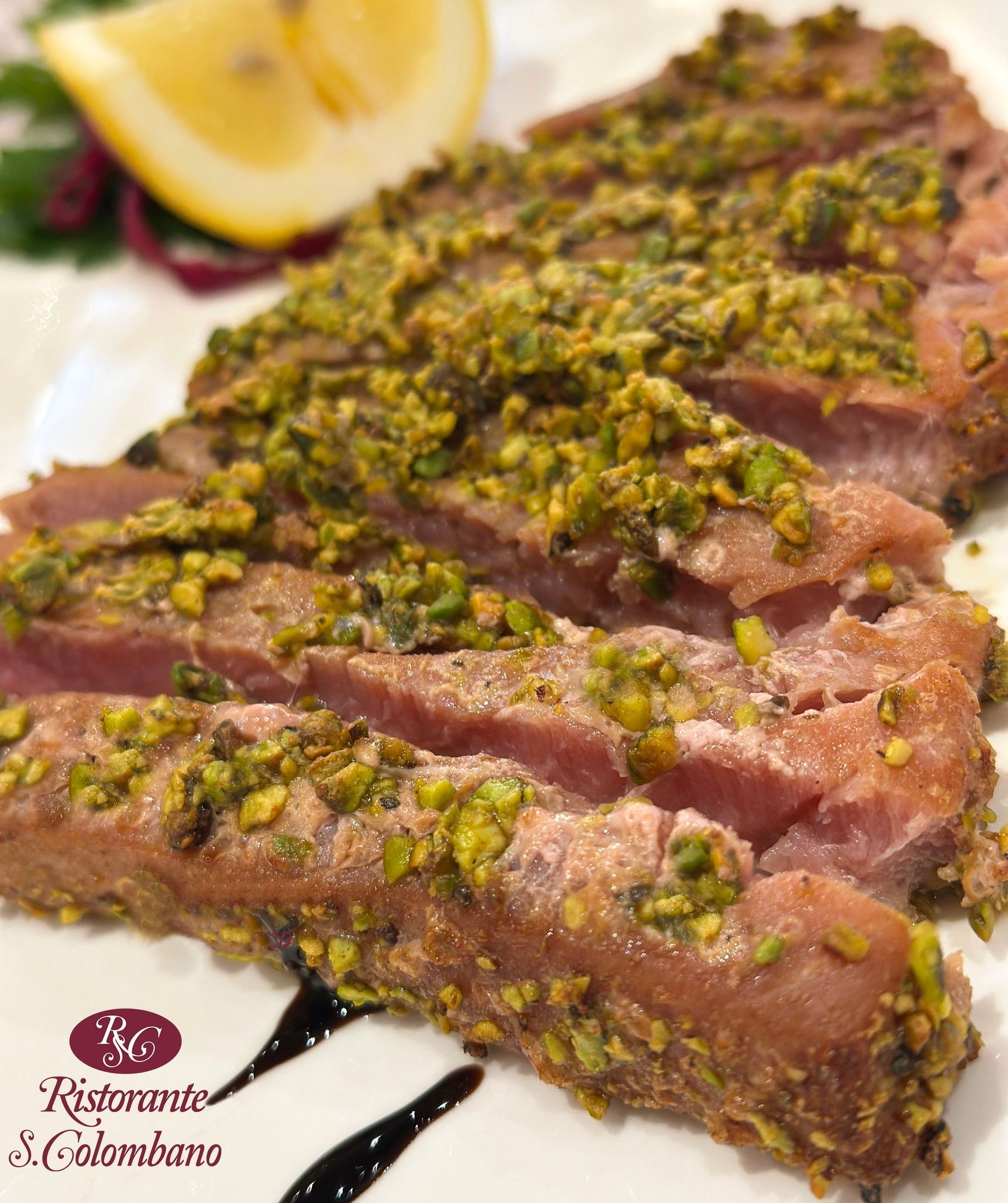 Una bistecca di tonno tagliata a fette e scottata in padella, ricoperta di pistacchi tritati, guarnita con uno spicchio di limone e un filo di aceto balsamico.