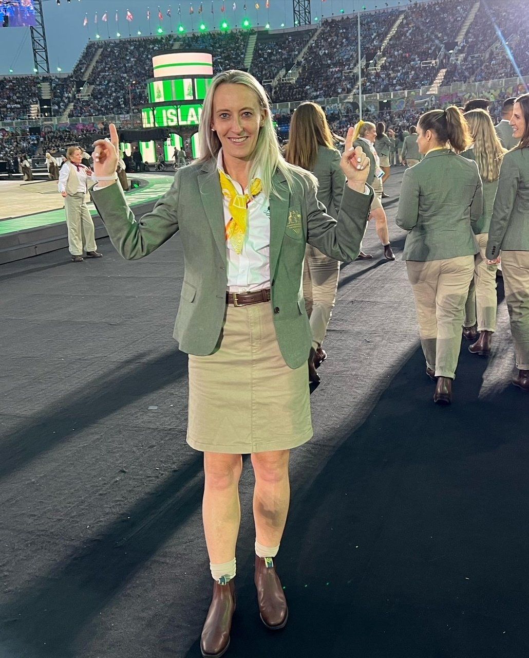 A woman in a green jacket and khaki skirt is standing in front of a crowd.