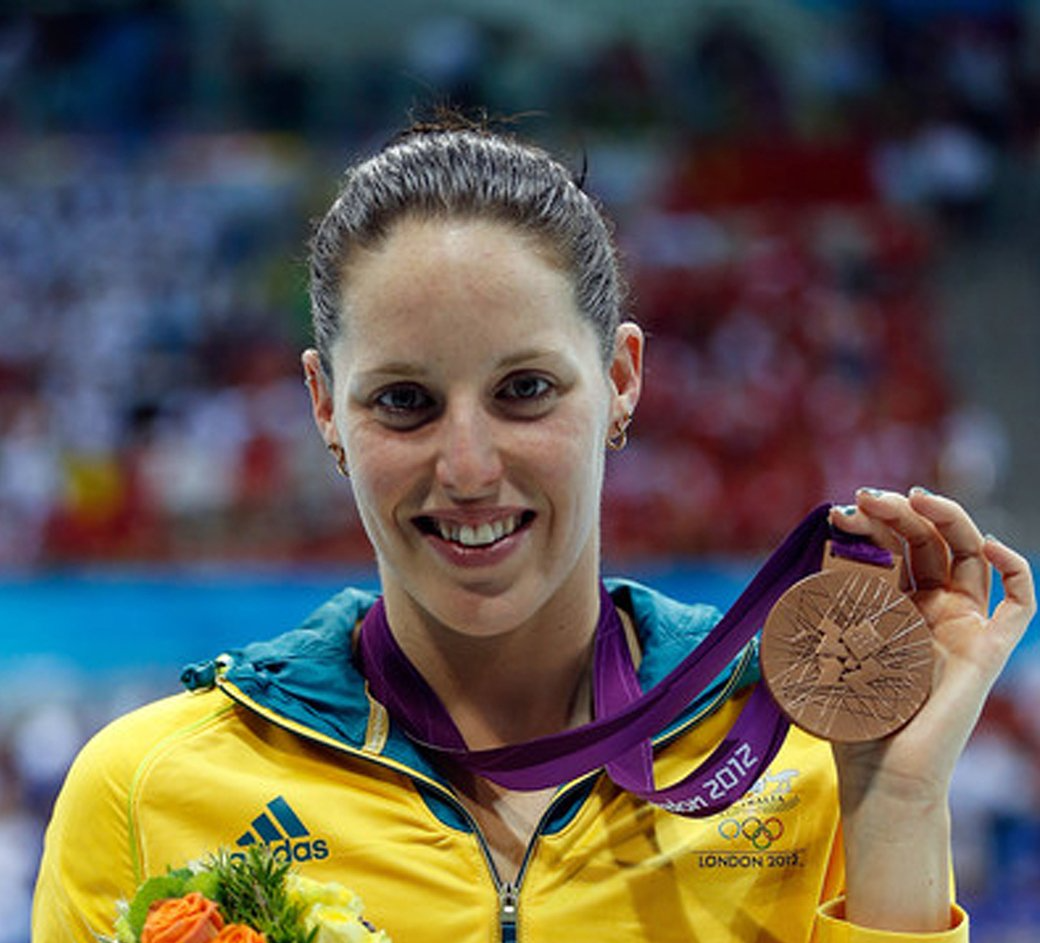 A woman in a yellow adidas jacket is holding a bronze medal
