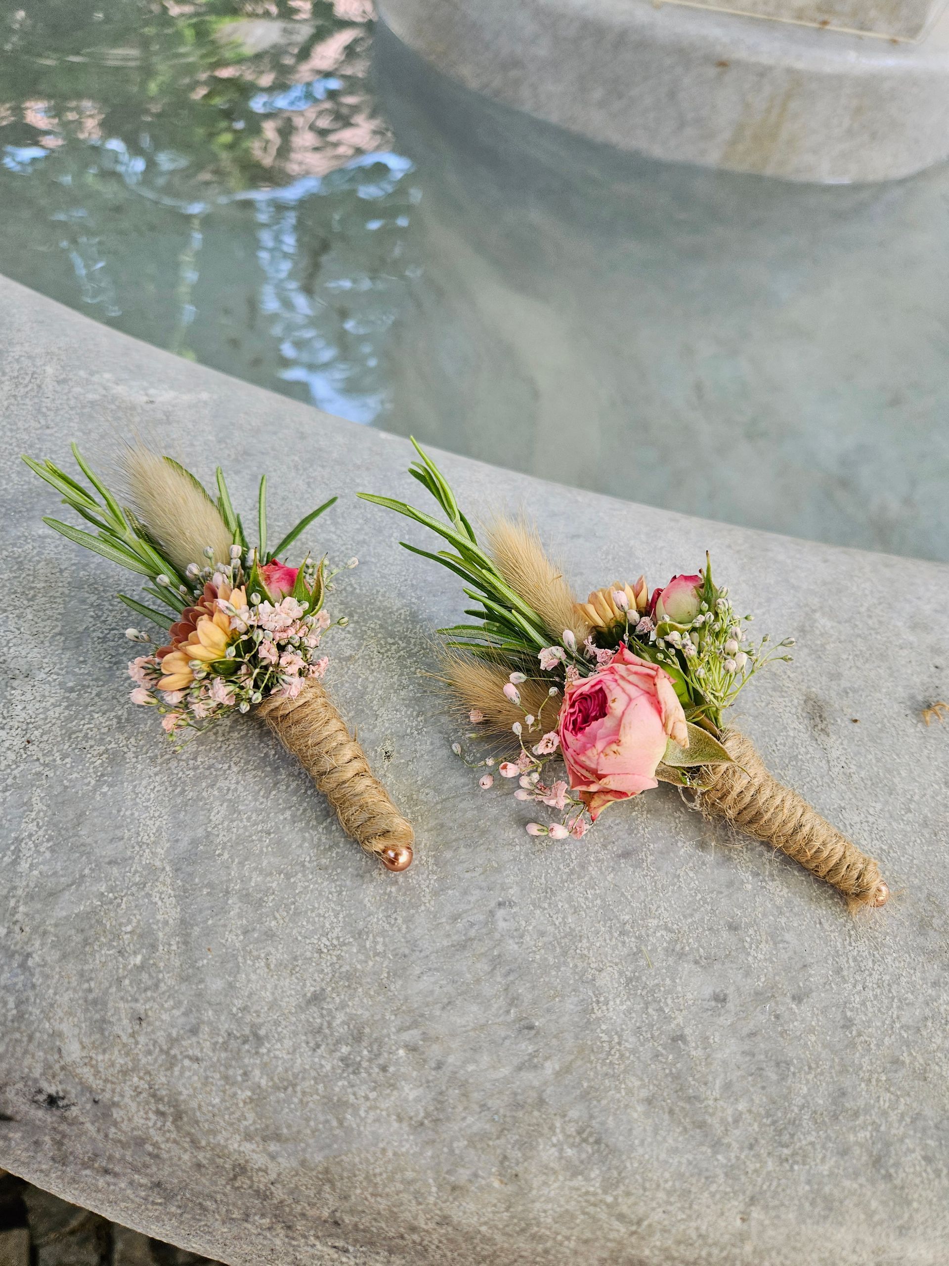 Zwei Boutonnieren mit Blumen stehen auf einer Marmorfläche neben einem Brunnen.